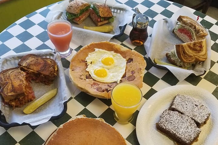A brunch spread on a green and white checked table: sandwiches in baskets, two eggs on a wide pancake, a syrup jug, orange juice and squares of banana bread.