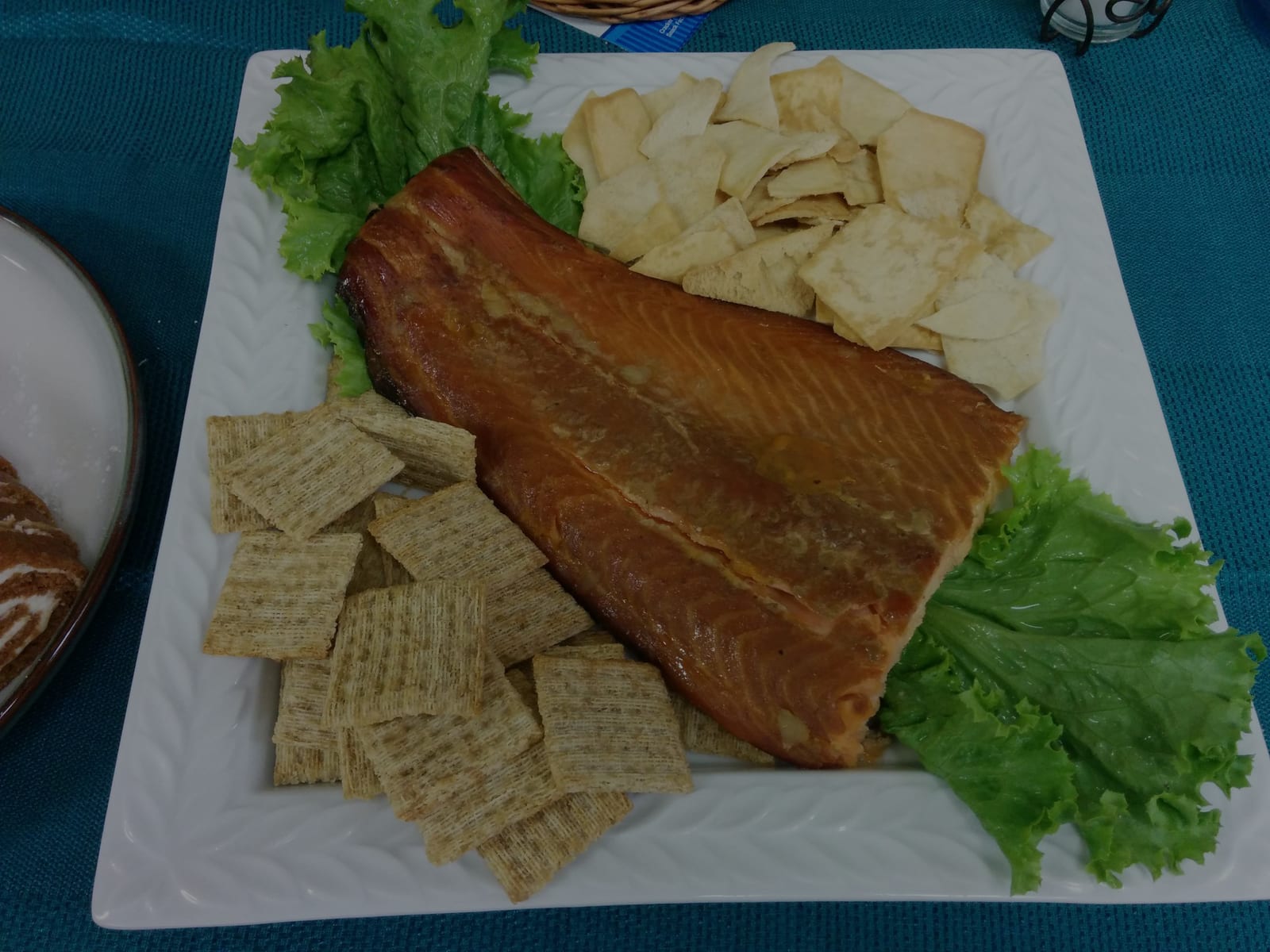 A whole apricot glazed smoked salmon fillet on a square white platter, ringed with crackers, pita chips and green leaf lettuce.