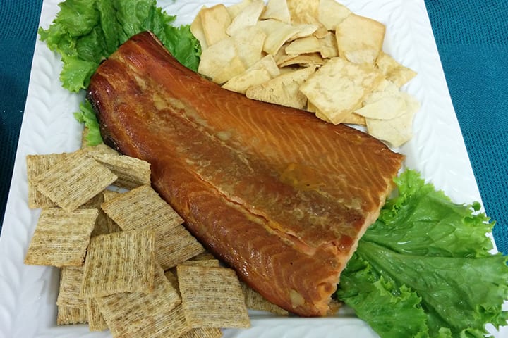 A fillet of apricot glazed smoked salmon on a white plate with wheat crackers, pita chips and lettuce.