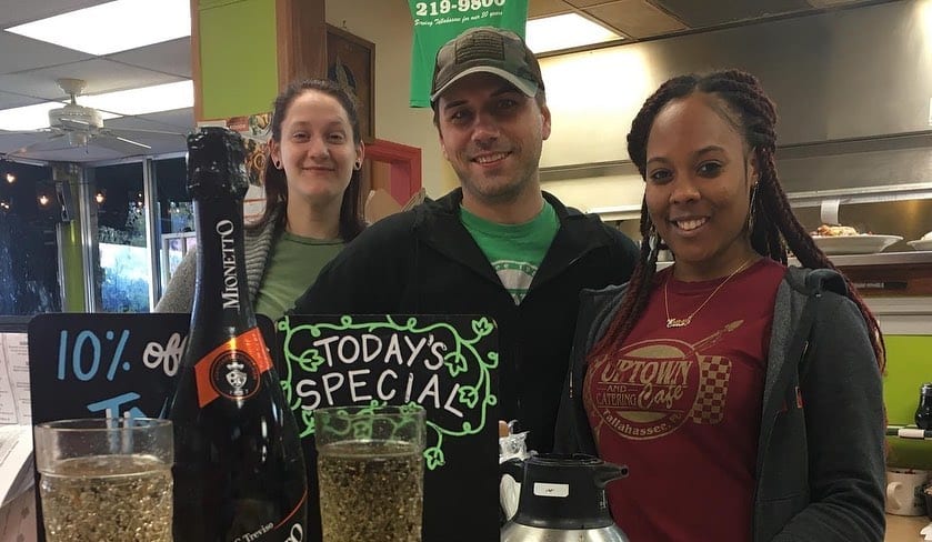 Three members of staff behind the counter beside a chalkboard reading Today’s Special, with a bottle of prosecco and two filled glasses.