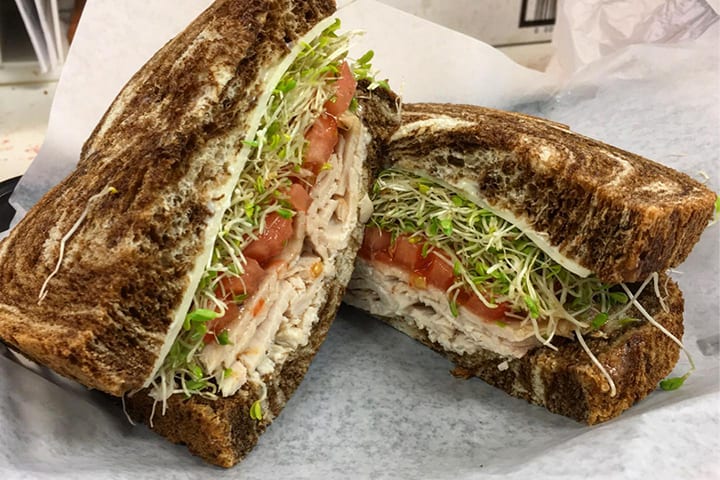 A turkey sandwich cut in half on toasted bread with tomato and sprouts, served with a pickle spear.