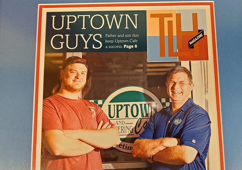 A copy of TLH Magazine with the cover line Uptown Guys, showing two men standing with their arms folded in front of the cafe’s window decal.